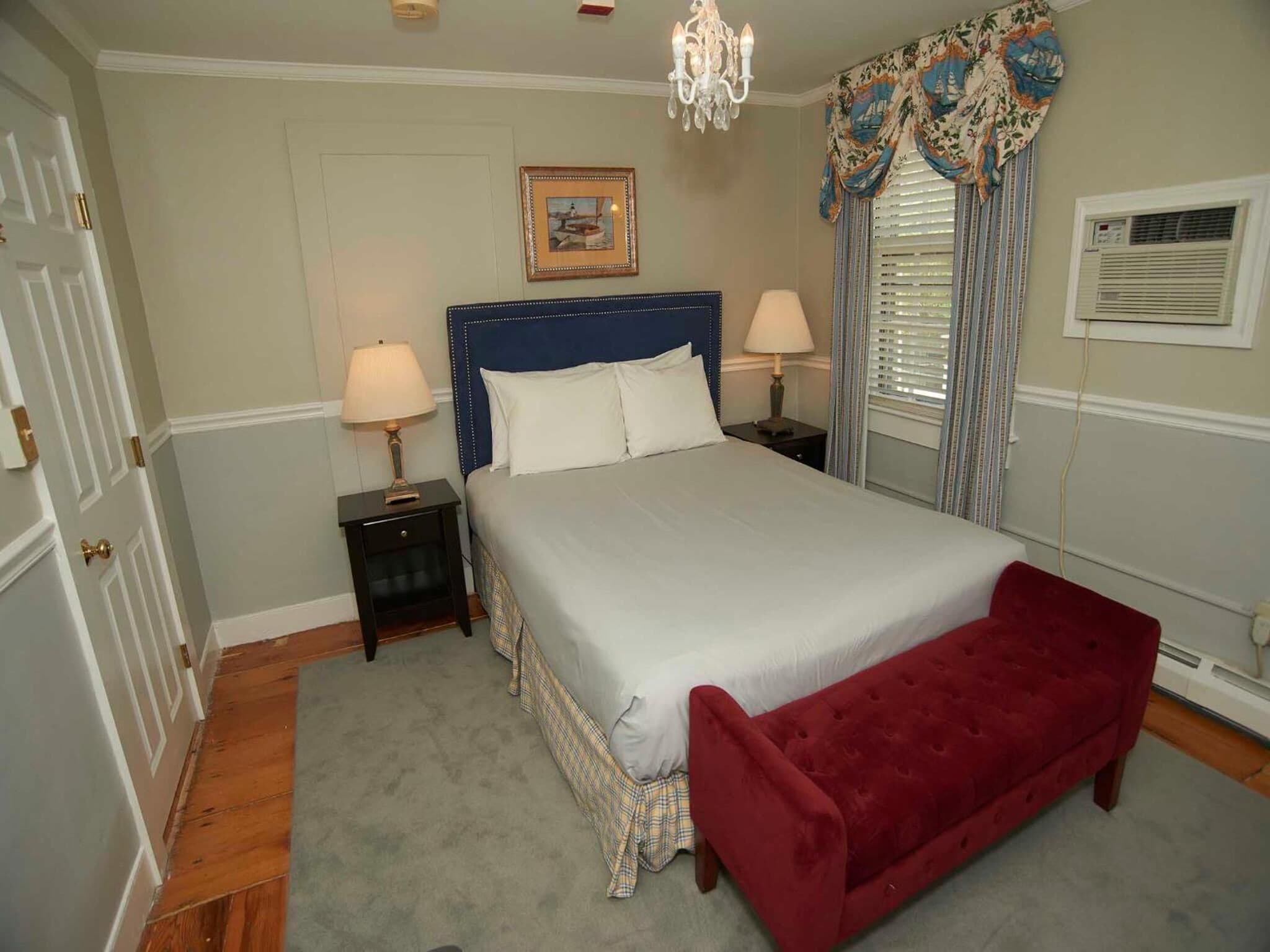 Une chambre à coucher bien aménagée, avec un grand lit, deux tables de chevet avec des lampes, un banc rouge, un lustre, un tableau encadré au mur et une fenêtre avec des rideaux.