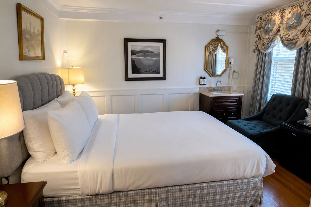 A hotel room with a large bed, a bedside table with a lamp, a chair, a vanity with a mirror, and a framed picture on the wall.