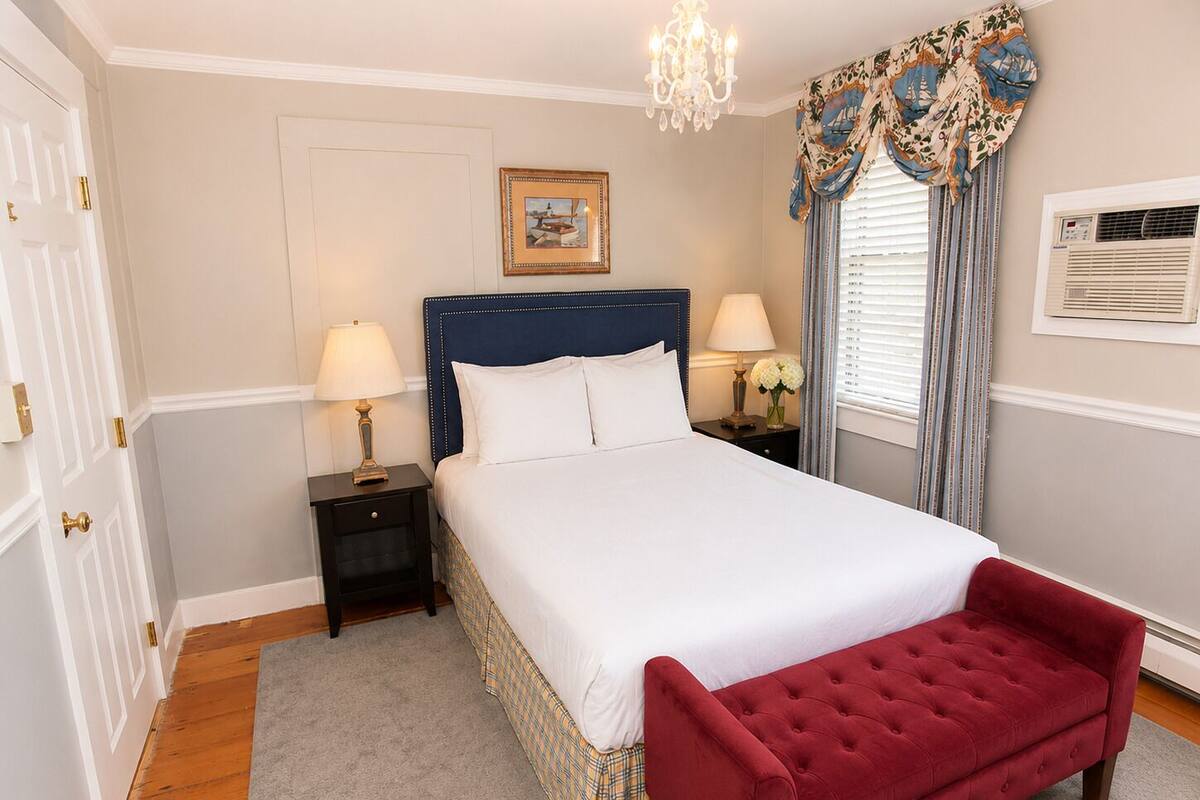 A bedroom with a bed, two bedside tables, a red bench, a chandelier, a window with curtains, a picture on the wall, and an air conditioning unit.