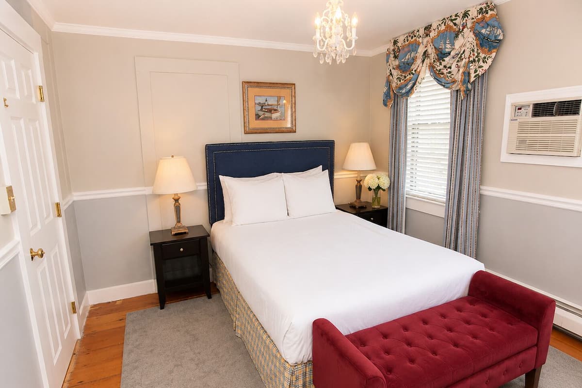 Quarto com cama, duas mesas de cabeceira, um banco vermelho, um candelabro, uma janela com cortinas, um quadro na parede e um ar condicionado.