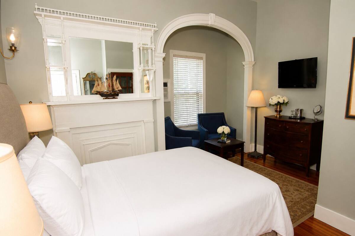 A bedroom with a bed, a chair, a dresser, a mirror, a television, and a window with blinds.