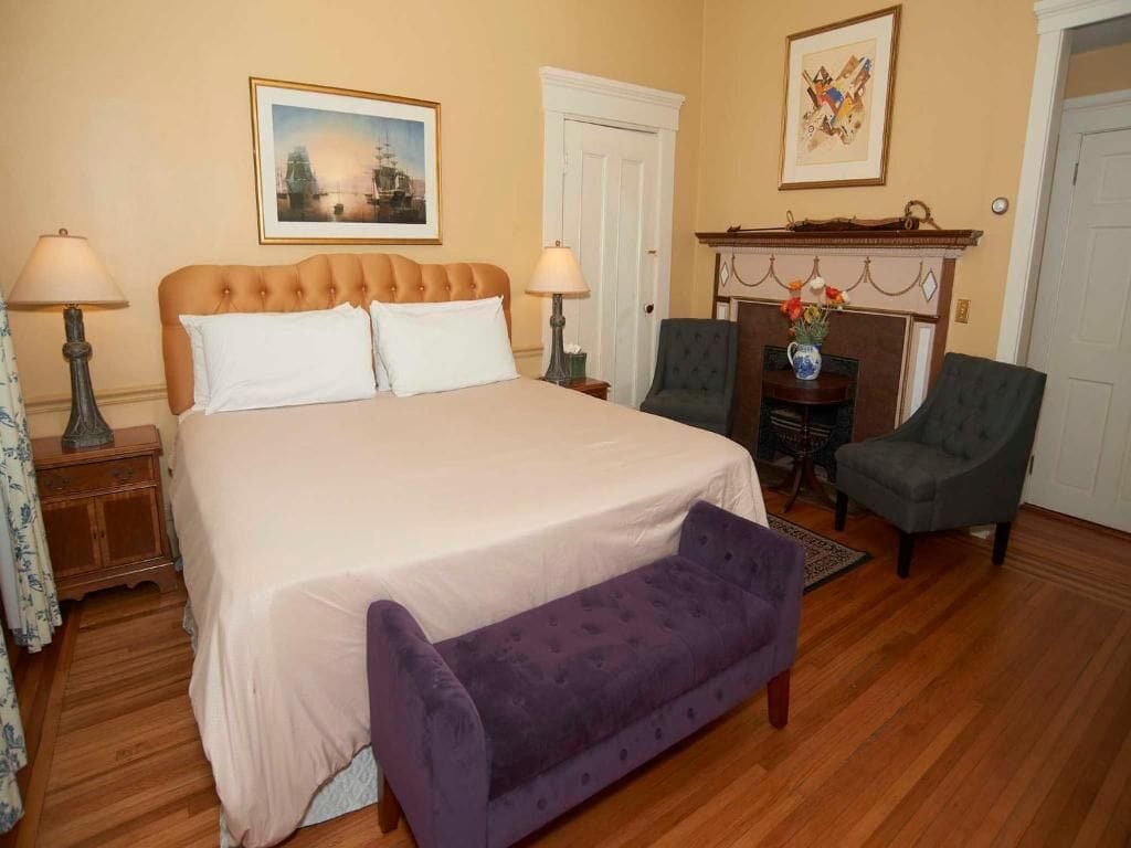 A bedroom with a large bed, two chairs, a bench, a fireplace, and two framed pictures on the wall.