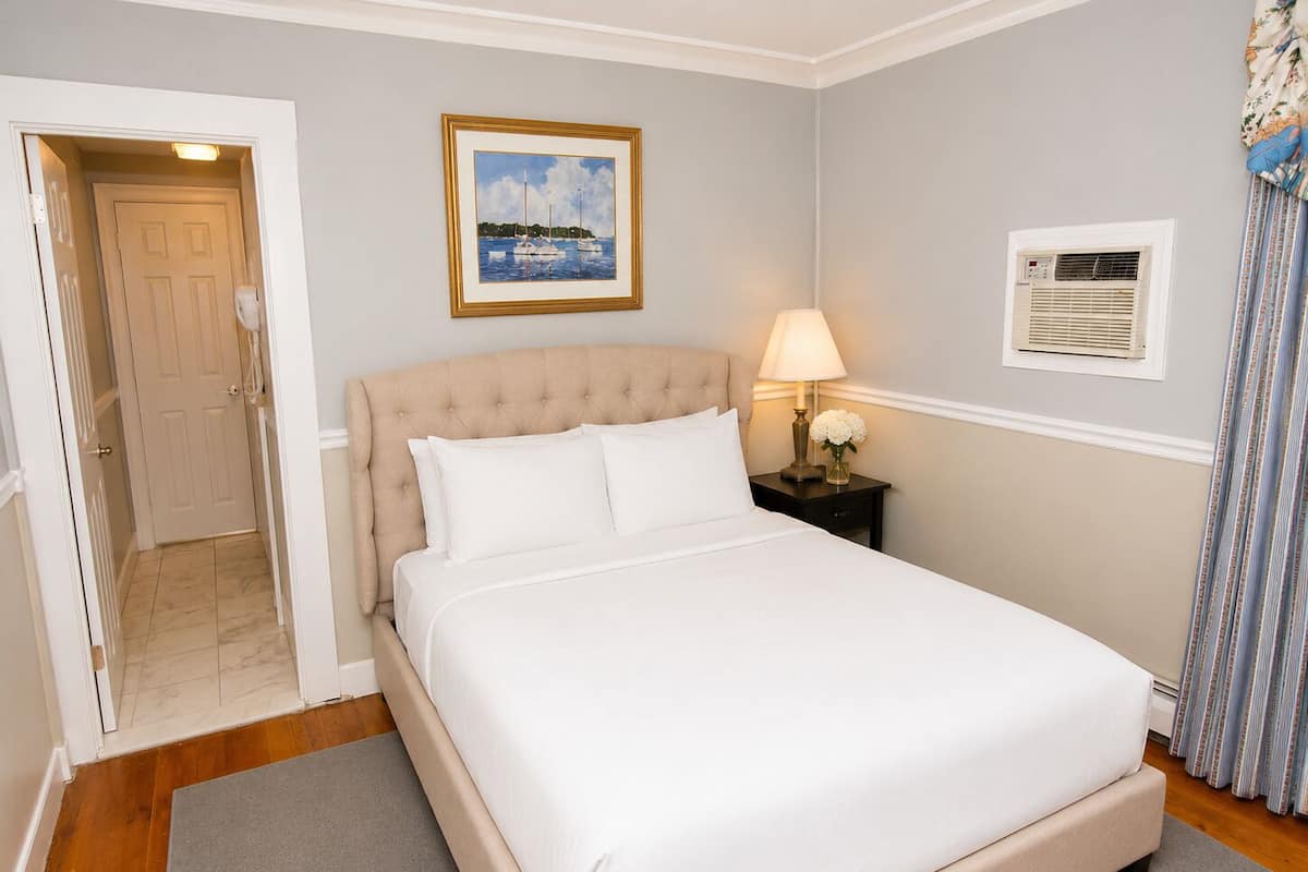 A neatly arranged bedroom with a bed, a bedside lamp, a framed picture on the wall, and a door leading to another room.