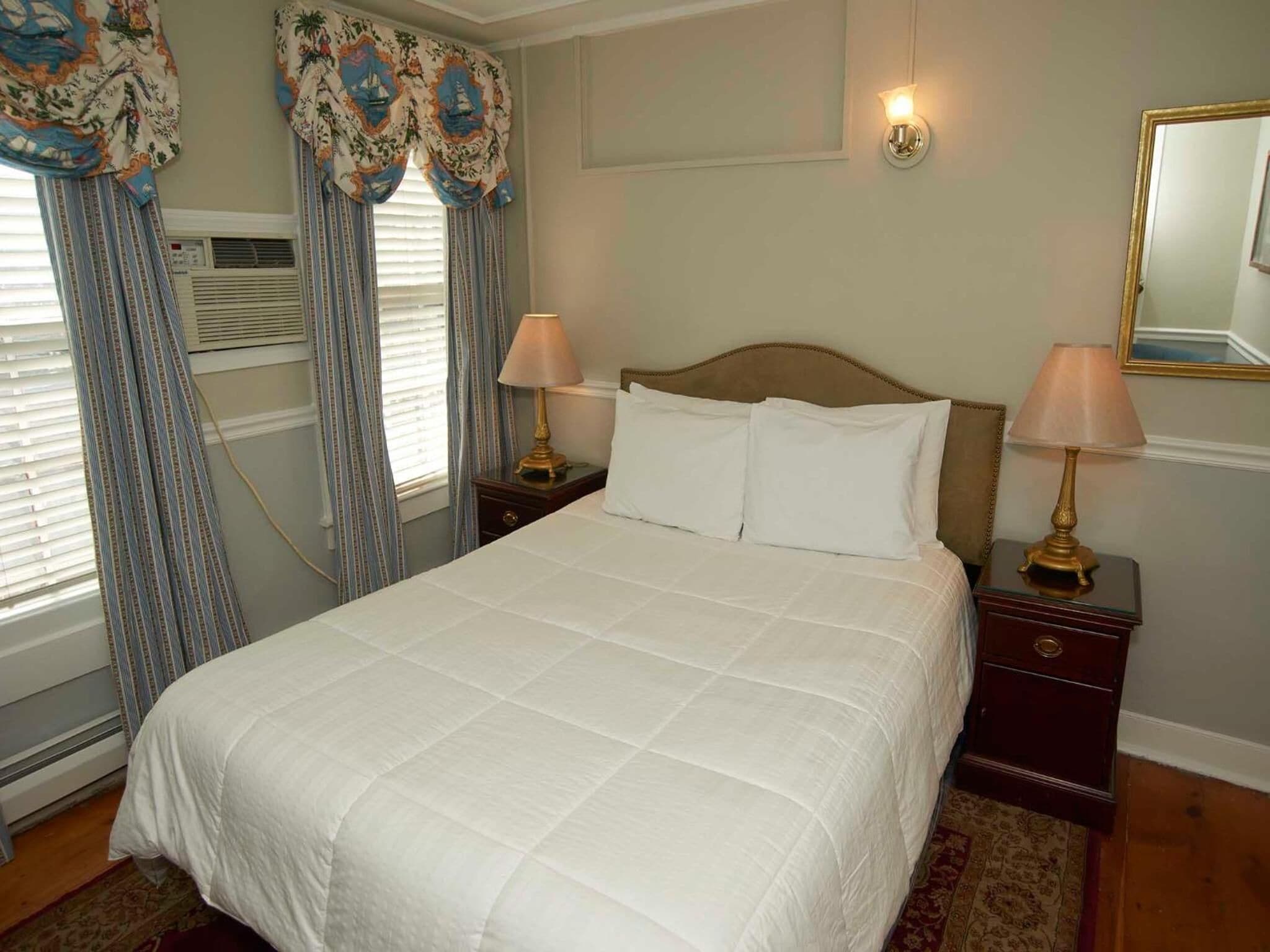 A neatly made bed with white bedding, two bedside lamps, and a window with curtains.