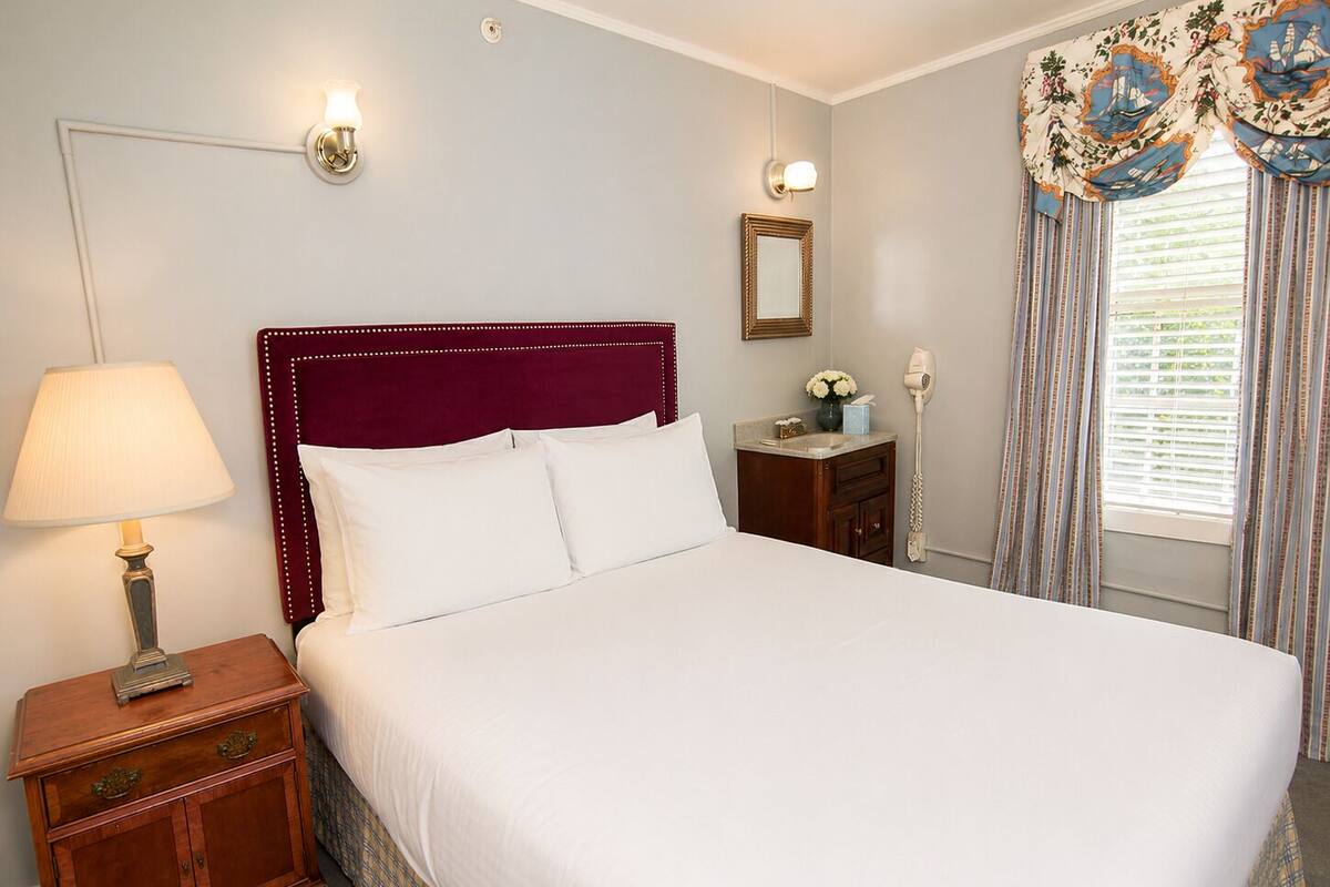 A neatly made bed with white linens, a wooden nightstand with a lamp, a small table with a vase of flowers, and a telephone on the wall.