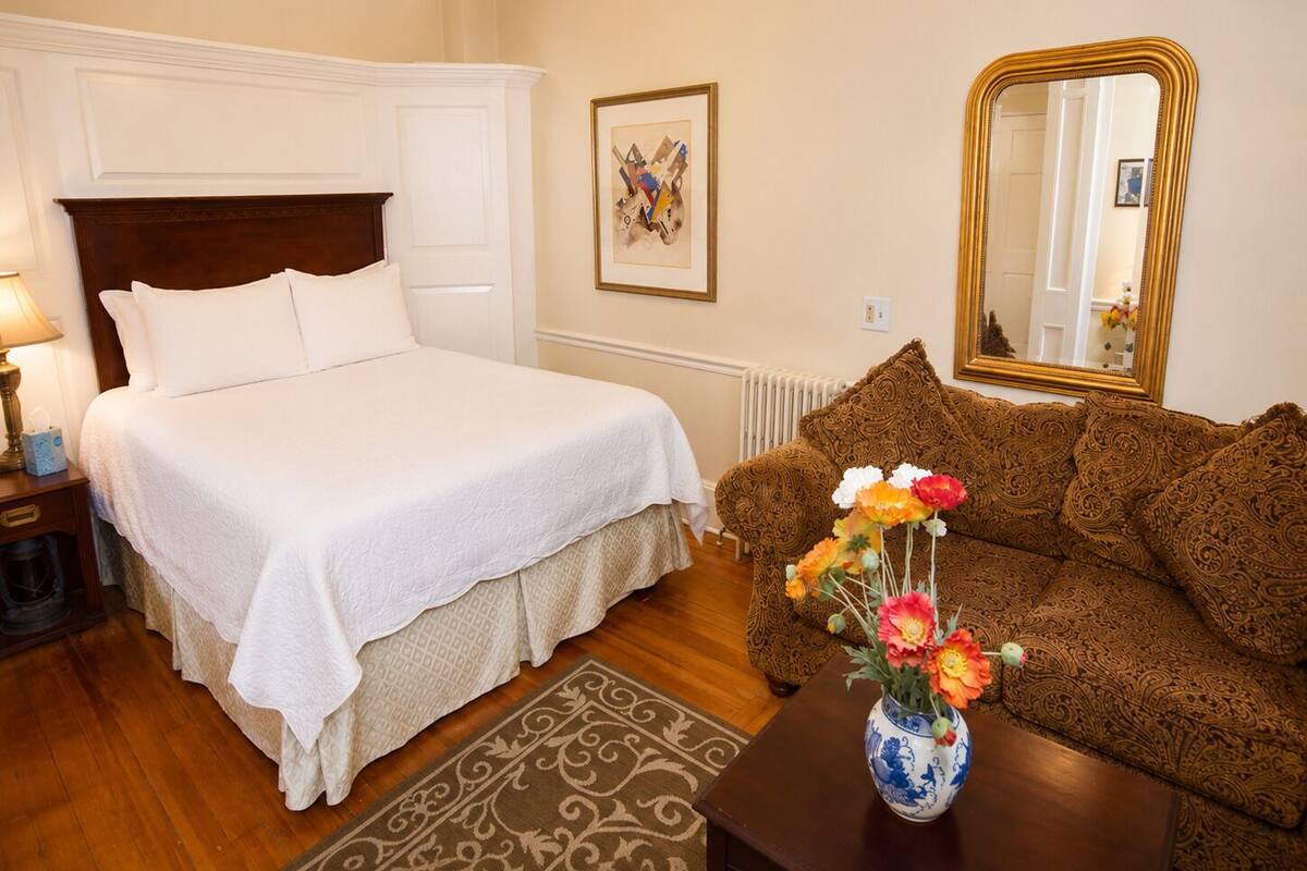 A hotel room with a bed, a sofa, a vase with flowers, a lamp, a framed picture, and a mirror.