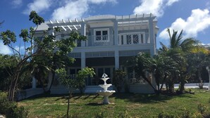 A two-story building with a white facade, a central entrance with a staircase, and a small fountain in the front yard. - Blue Orchid Villa (Moss Town)