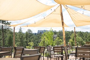 An outdoor seating area with wooden tables and chairs under a large canopy, surrounded by trees and a mountainous backdrop. - Under Canvas Mount Rushmore (Keystone)
