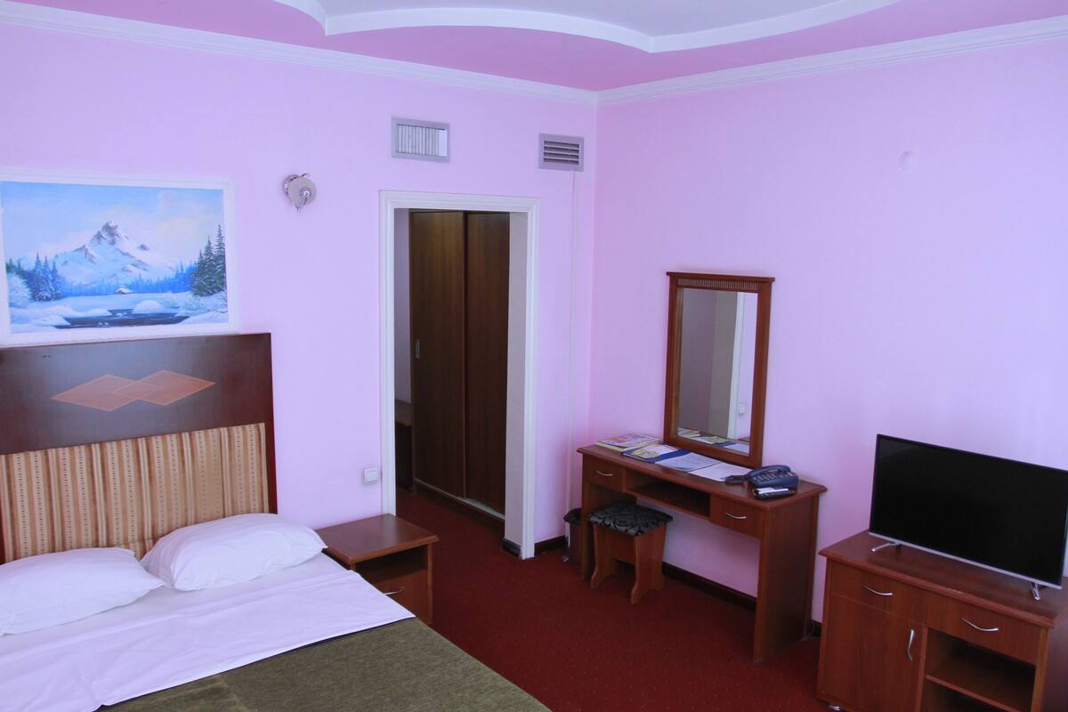 Ein Hotelzimmer mit Bett, Schreibtisch mit Spiegel, Fernseher und einem Bild an der Wand.