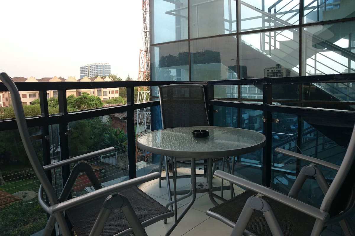 Un balcon doté d’une table et de chaises en verre, offrant une vue sur un paysage urbain avec ses bâtiments et sa verdure.