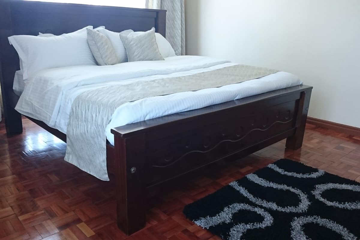 Un lit bien fait, avec du linge de lit blanc et une tête de lit sombre, un plancher en bois et un tapis à motifs noir et blanc.