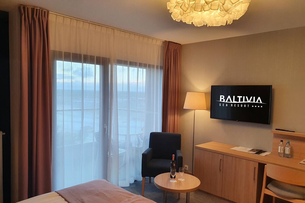 Hotellihuone, jossa on sänky, työpöytä tuolilla, televisio ja suuret ikkunat, joista avautuu näkymä merelle.