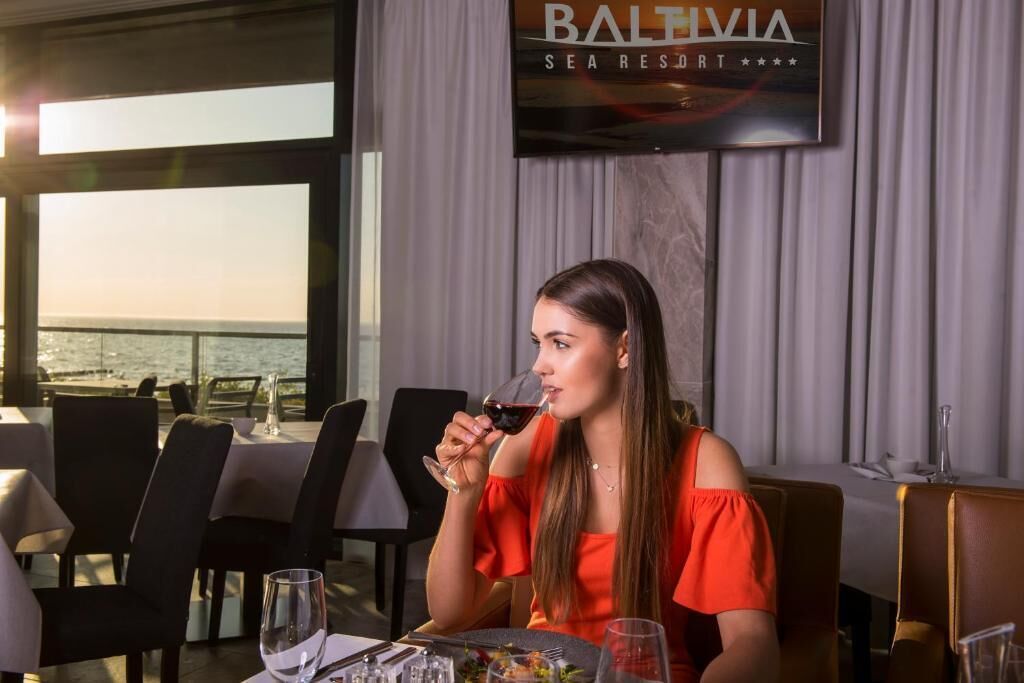 A woman seated at a table in a restaurant with a view of the sea, a sign reading "Baltivia Sea Resort", and a glass of red wine on the table.