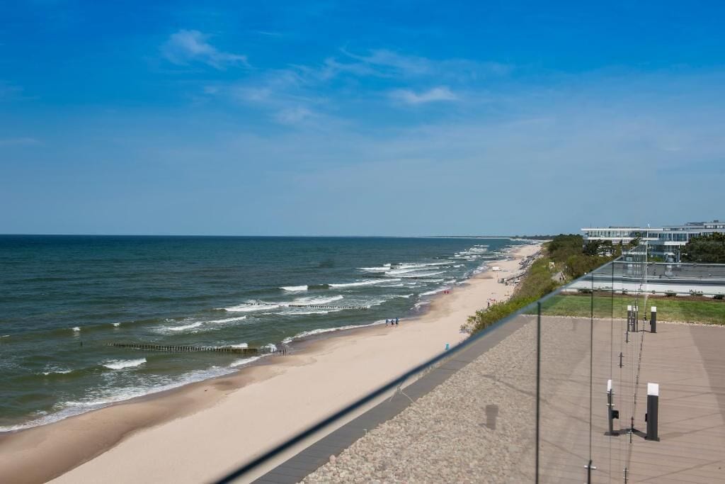 A coastal walkway with a beach and ocean view, a paved path, and a building in the background.
