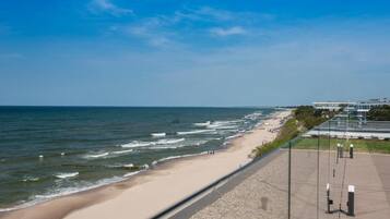 En strandpromenad längs kusten med utsikt över stranden och havet, en asfalterad gångväg och en byggnad i bakgrunden.
