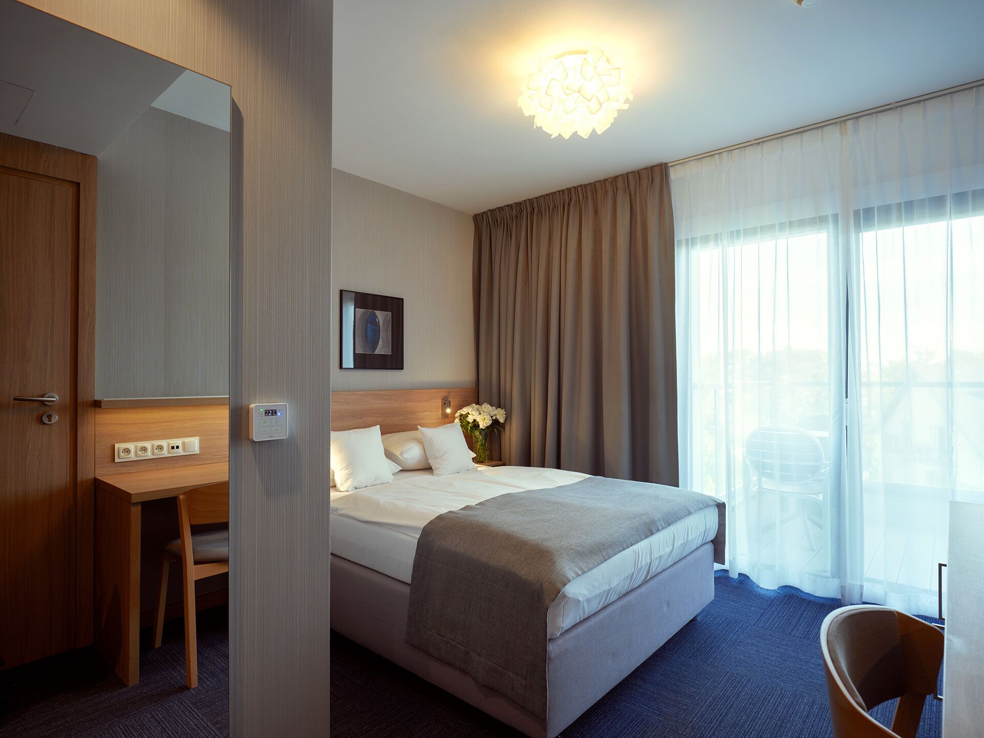 A hotel room with a bed, a desk, a chair, a bedside table, a lamp, and a window with curtains.