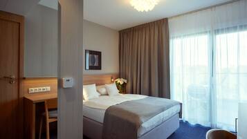 A hotel room with a bed, a desk, a chair, a bedside table, a lamp, and a window with curtains.