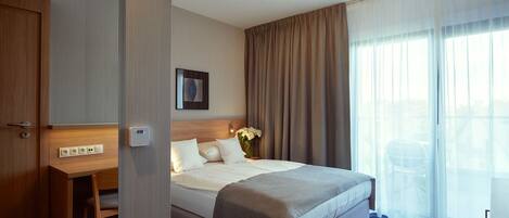 A hotel room with a bed, a desk, a chair, a bedside table, a lamp, and a window with curtains.
