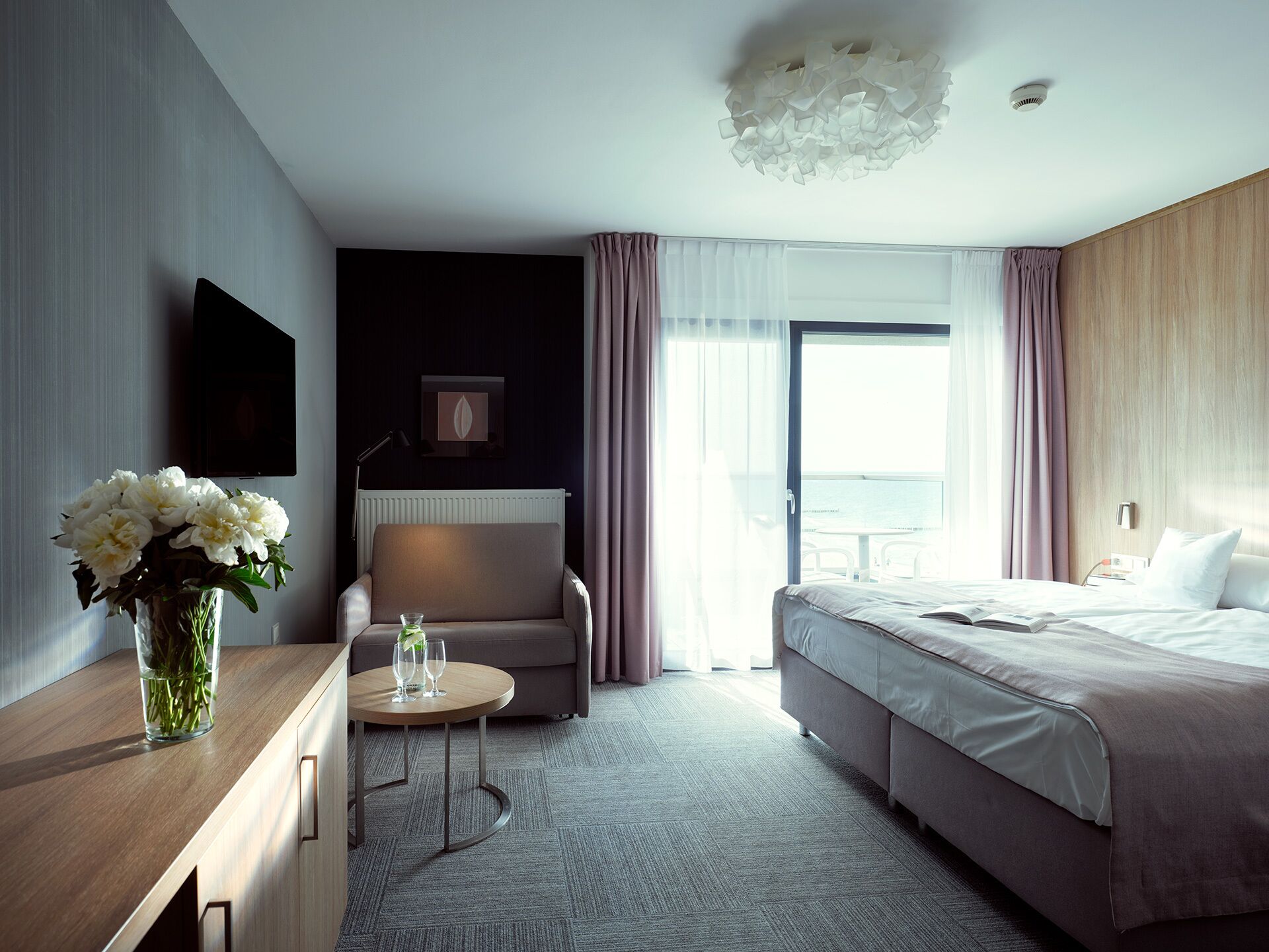 A modern hotel room with a bed, a seating area with a table, a TV, and a view of the outdoors.