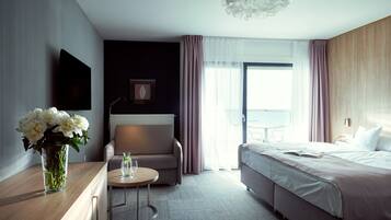 A modern hotel room with a bed, a seating area with a table, a TV, and a view of the outdoors.