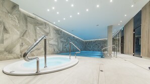 Un espace piscine intérieure moderne, avec un mur en marbre, une piscine à l’eau bleutée et un escalier en métal.