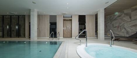 Une grande piscine intérieure, dotée d’un espace séparé avec un jacuzzi, le tout agrémenté d’éléments architecturaux modernes et d’un mur en marbre.