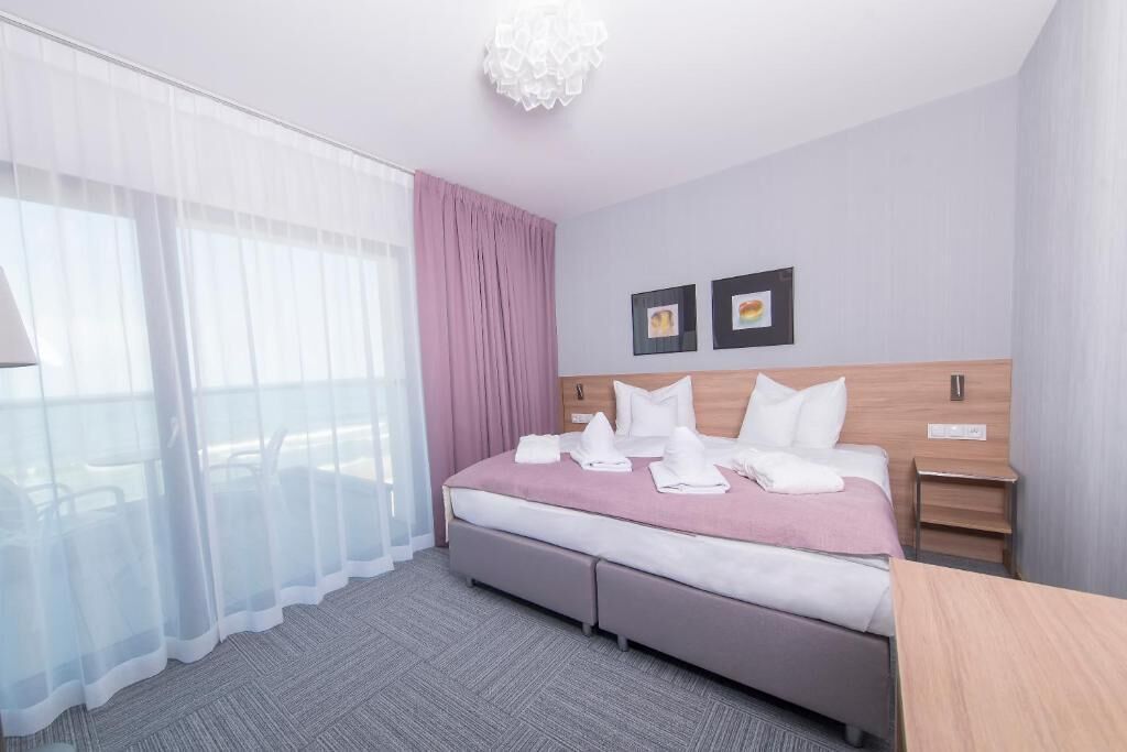 A hotel room with a bed, a bedside table, a nightstand, a window with sheer curtains, and a wall-mounted light fixture.
