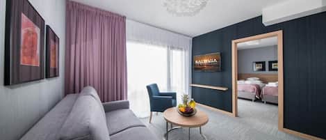 Una habitación de hotel moderna con un sofá gris, una mesa redonda de centro con fruta, un televisor y una cama con cabecero de madera.