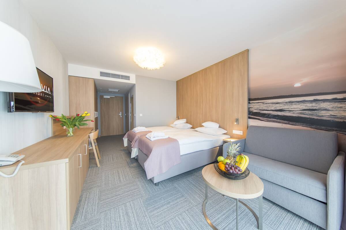Moderni hotellihuone, jossa on sänky, sohva, kirjoituspöytä puhelimineen, pieni pöytä hedelmillä ja seinälle asennettu televisio.
