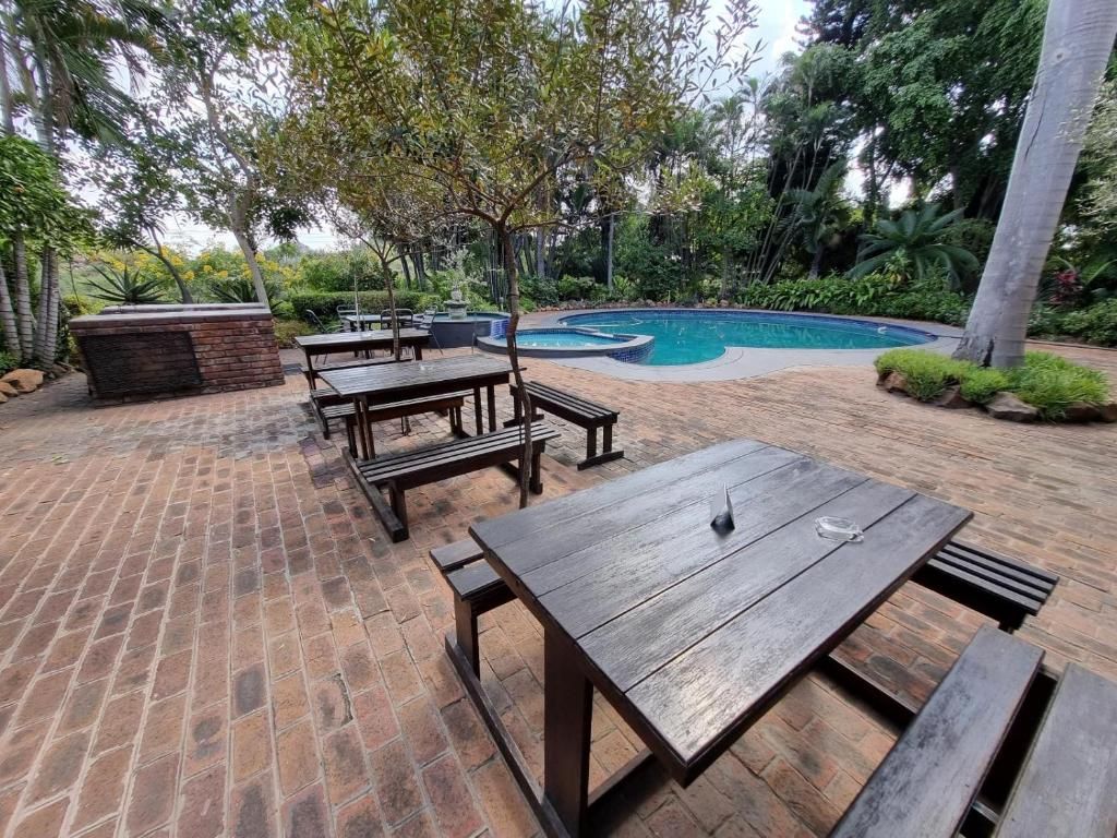 Un patio al aire libre con mesas y bancos de madera, suelo de ladrillo y una piscina rodeada de vegetación.