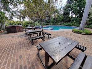Um pátio externo com mesas e bancos de madeira, piso de tijolos e uma piscina rodeada por vegetação.