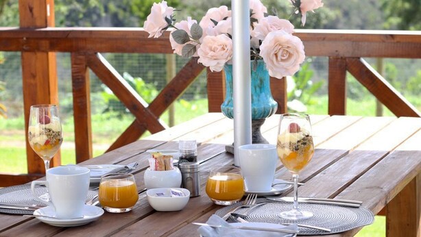 Une table en bois, dressée pour le petit-déjeuner, avec des tasses blanches, un verre de jus d’orange et un vase rempli de fleurs roses.