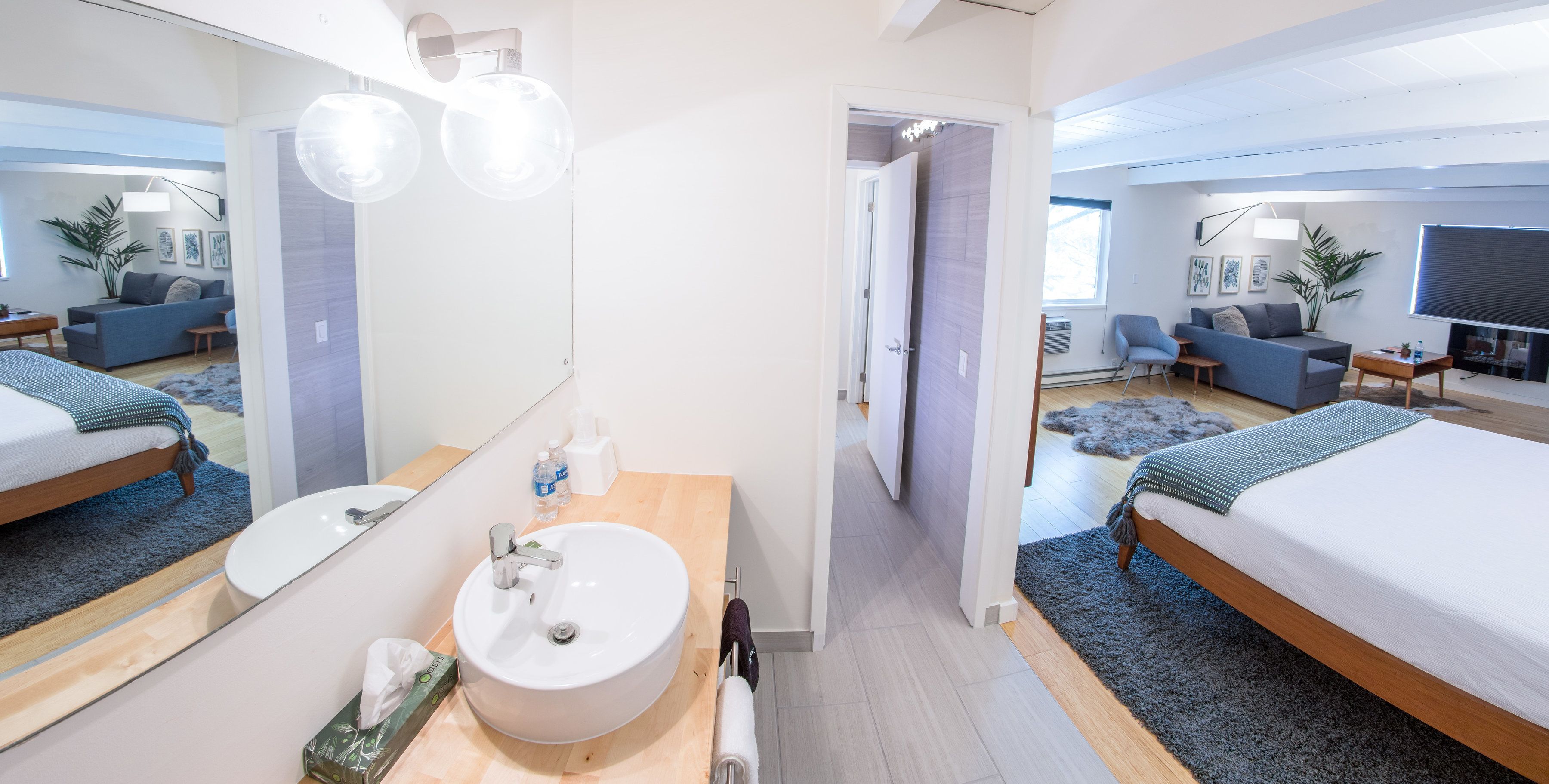 Et moderne hotelværelse med en stor, rund vask, et spejl, en seng med et gråt tæppe, et lille bord og et fjernsyn.