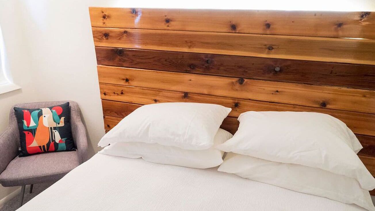 Una cama con ropa blanca y un cabecero de madera, un sillón gris con un cojín colorido, y una ventana con cortinas blancas.