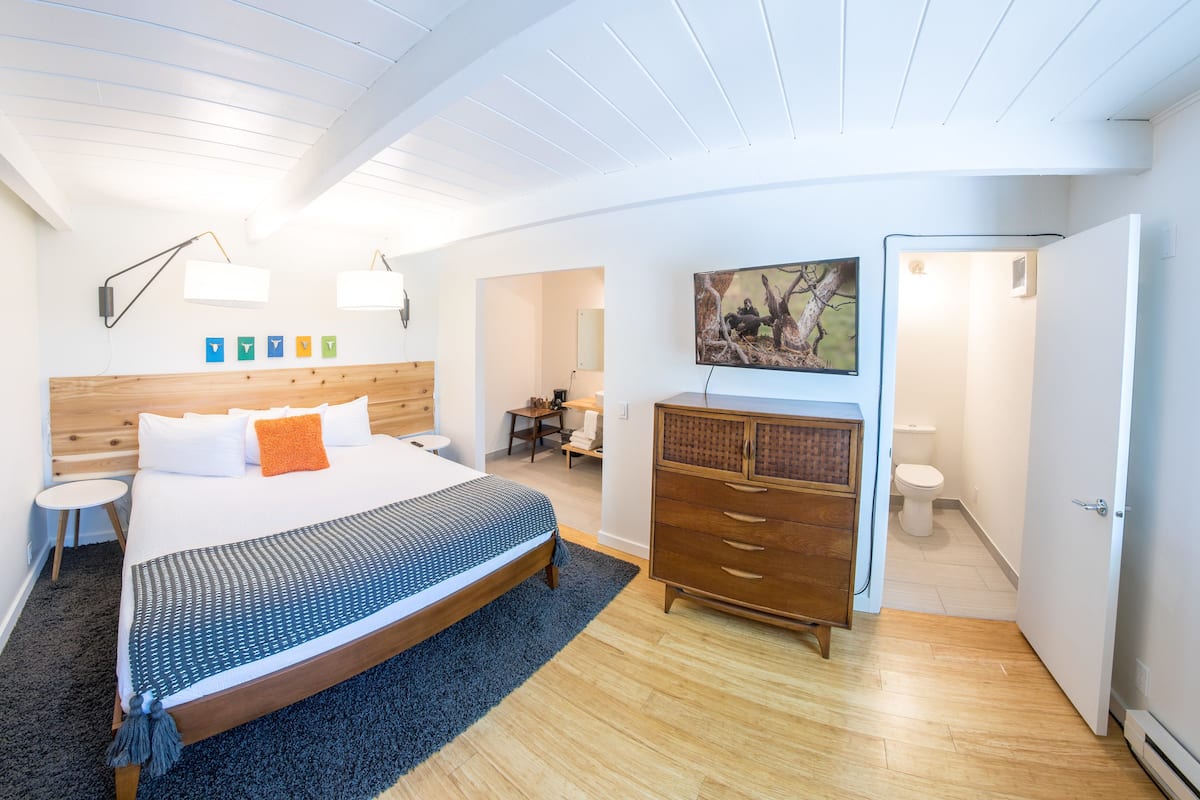 A modern bedroom with a wooden headboard, a large bed, a dresser, a desk, and a bathroom with a toilet and shower.