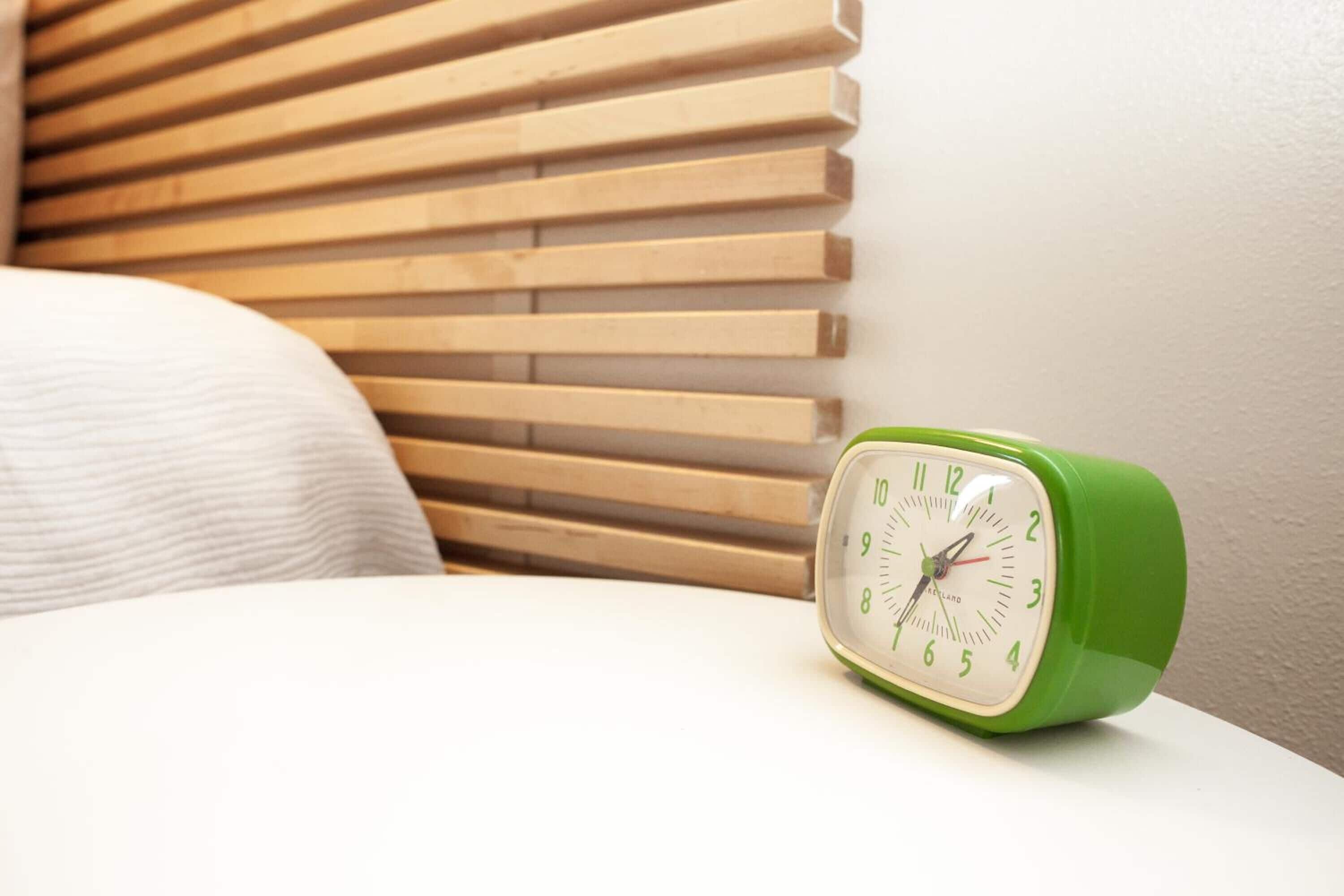 A green alarm clock on a white bedside table with a wooden slat headboard in the background.