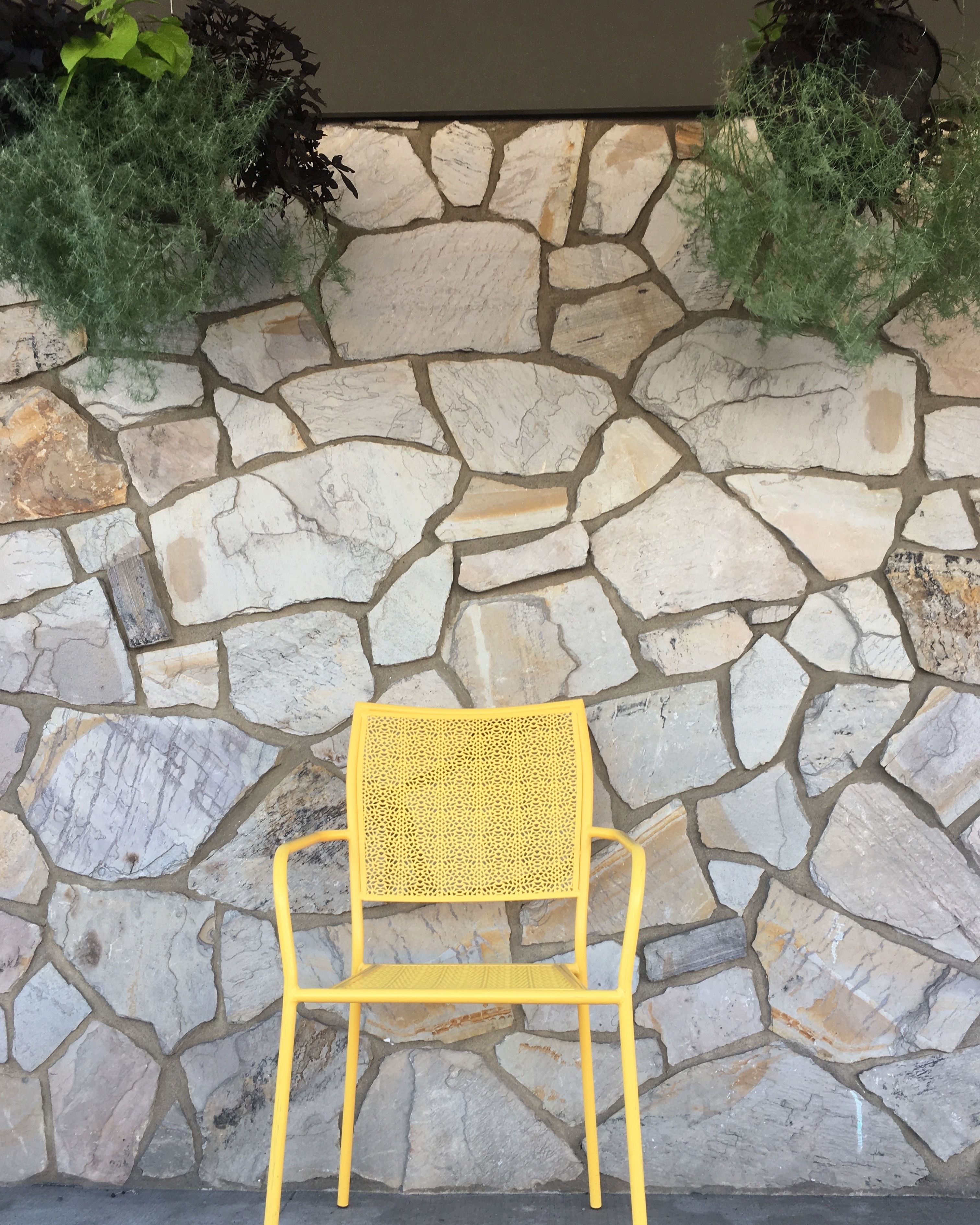 Una silla amarilla frente a un muro de piedra con plantas encima.