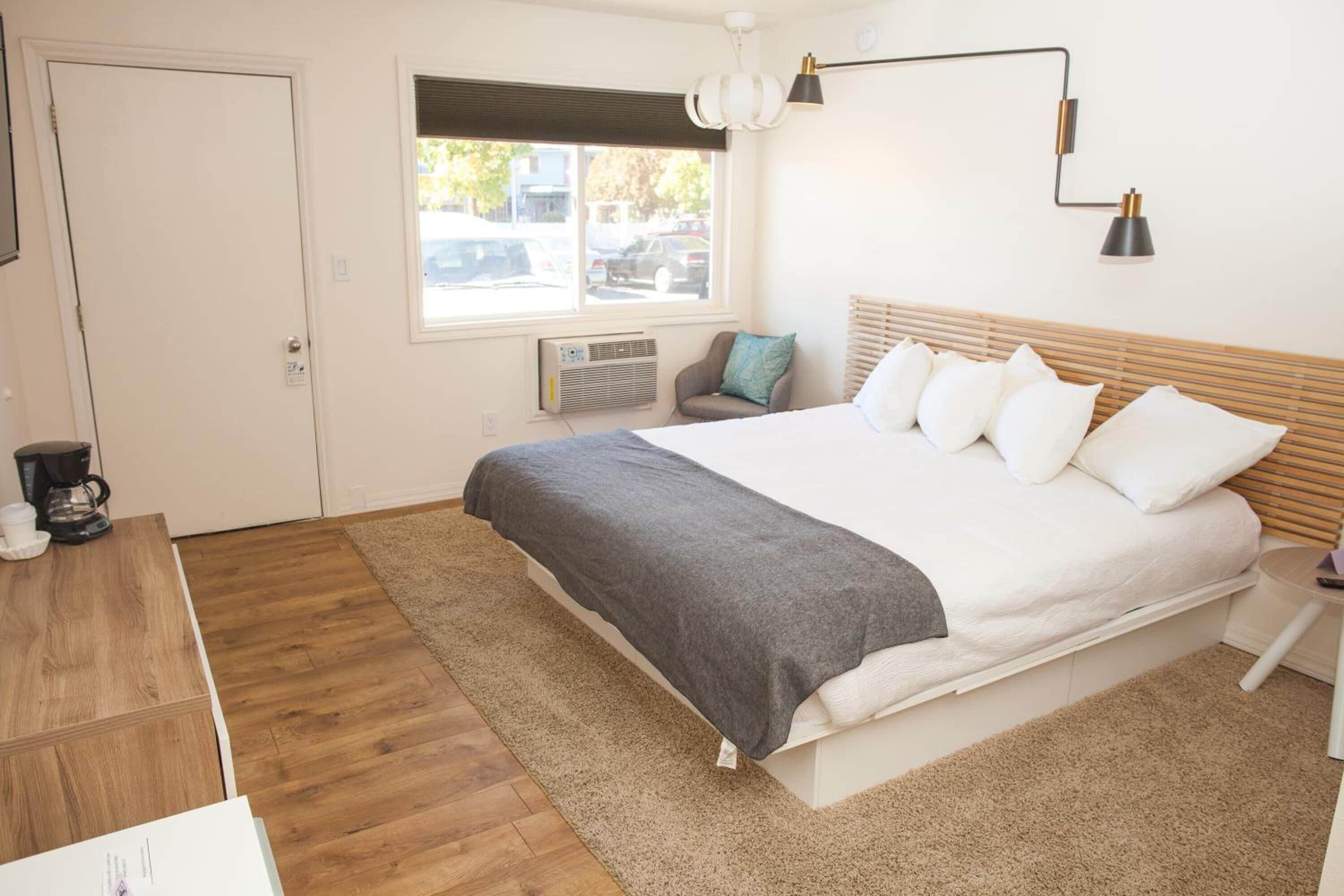 A bedroom with a bed, a coffee maker, a window, and a wall-mounted lamp.