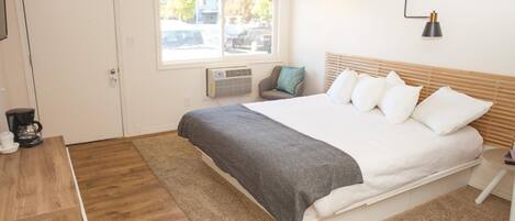A bedroom with a bed, a coffee maker, a window, and a wall-mounted lamp.