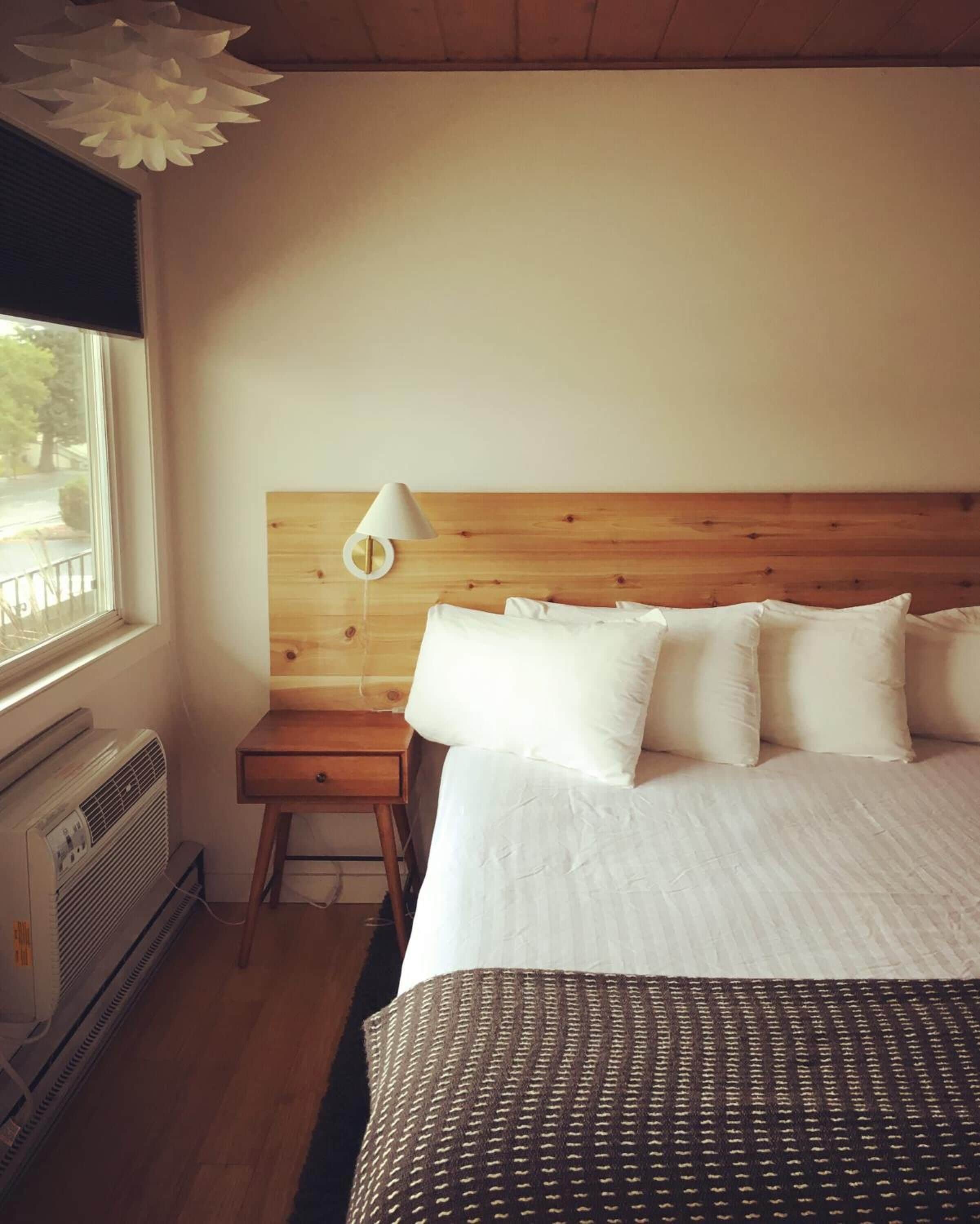 Een slaapkamer met een houten hoofdbord, wit beddengoed, een nachtkastje met een lamp en een raam met jaloezieën.