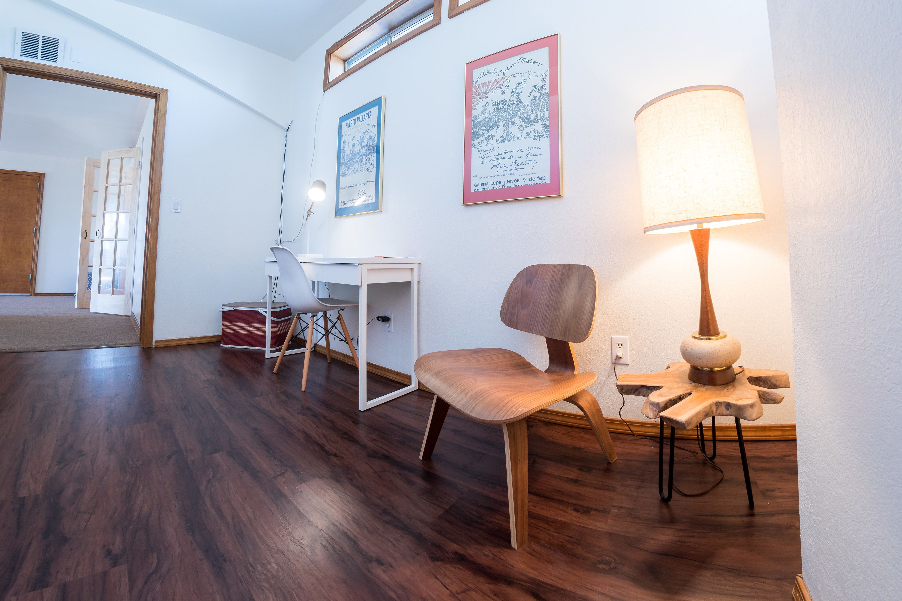 A room with a wooden floor, a desk with a chair, a lamp, and two framed pictures on the wall.