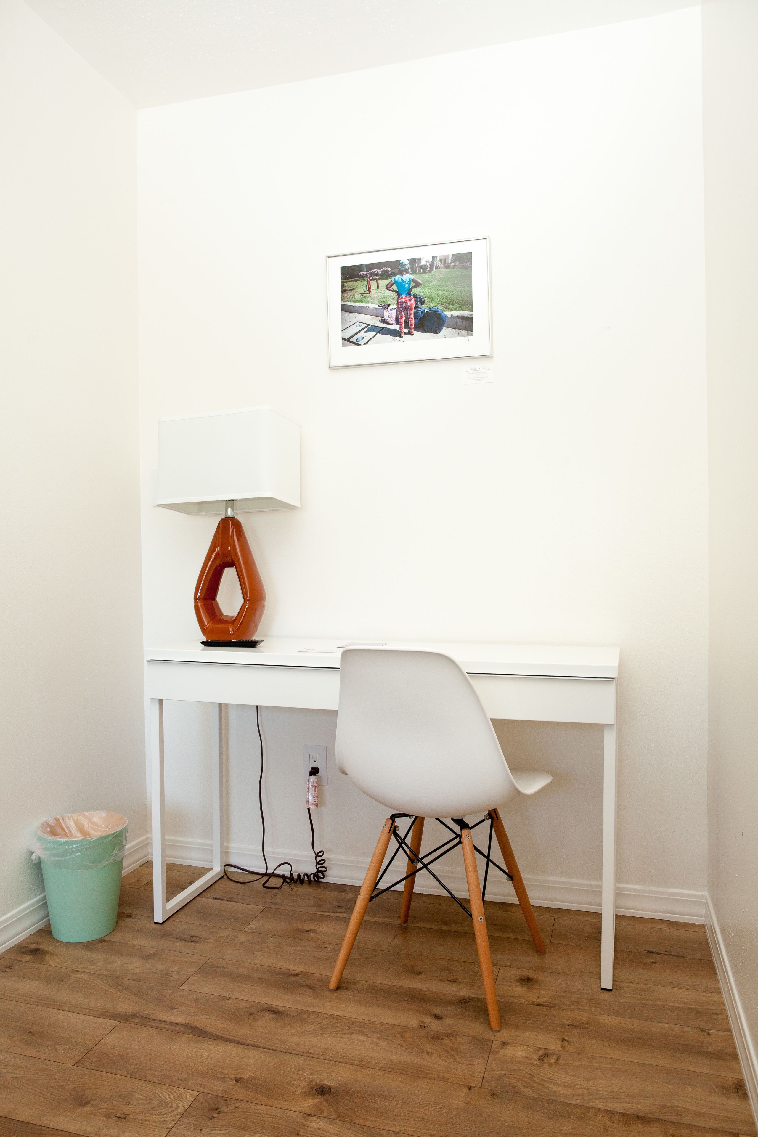 Un bureau blanc avec une lampe au design moderne, un tableau encadré accroché au mur et une chaise blanche aux pieds en bois.
