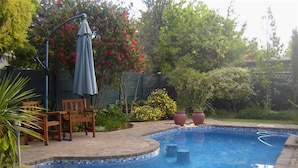 A swimming pool with a tiled edge, surrounded by a brick patio, wooden chairs, and a blue umbrella. - La Boheme Guesthouse (Bloemfontein)