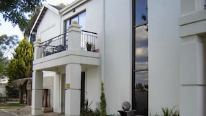 A modern two-story building with a balcony, a garden area with plants and a water feature, and a clear sky. - La Boheme Guesthouse (Bloemfontein)