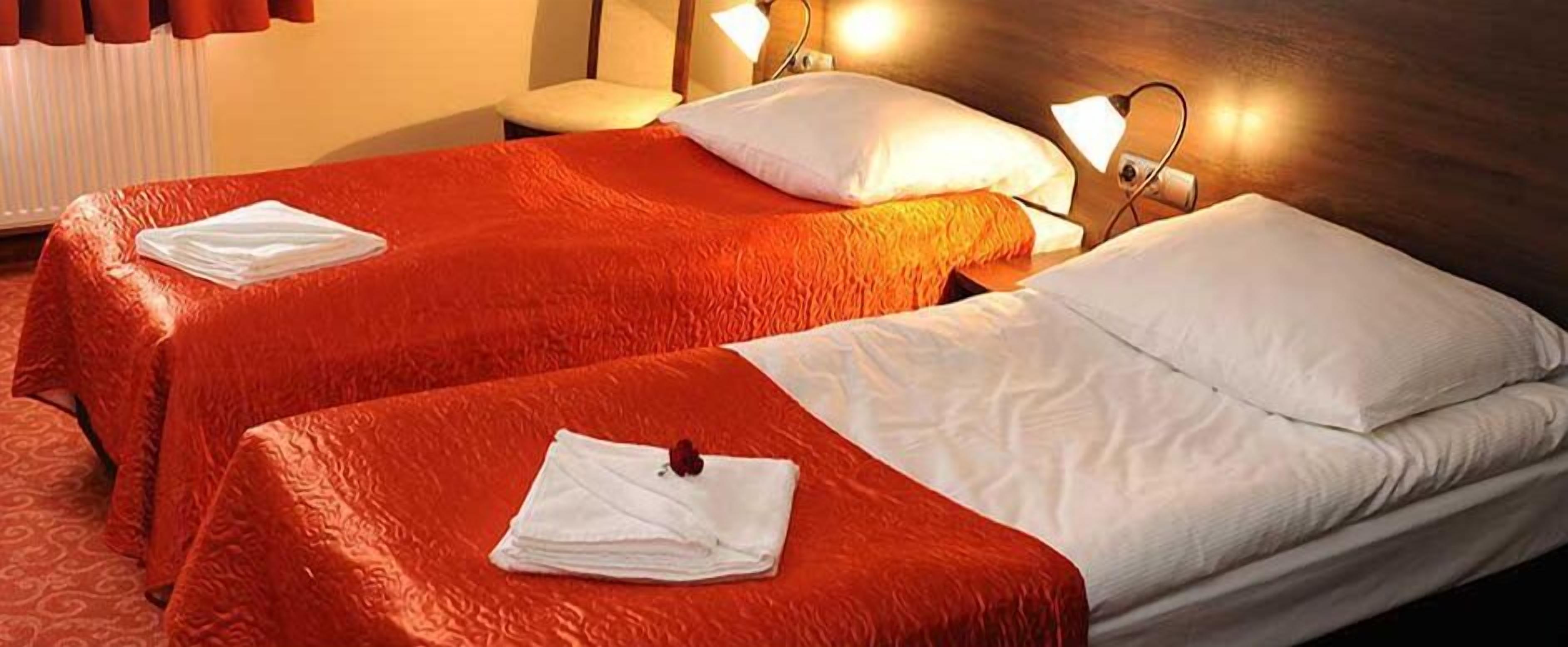Habitación de hotel con dos camas, cada una con colcha roja y almohadas blancas. Hay dos lámparas de noche y una mesita con una lámpara.