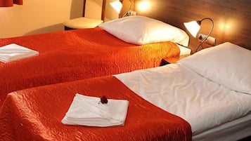 Habitación de hotel con dos camas, cada una con colcha roja y almohadas blancas. Hay dos lámparas de noche y una mesita con una lámpara.