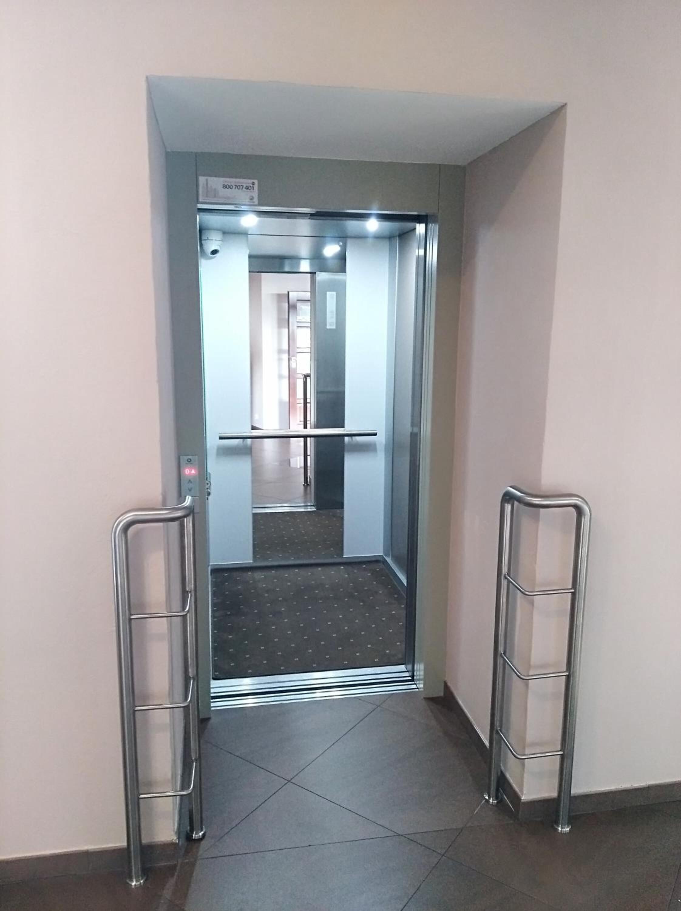 An elevator with a metal handrail, a control panel with a button, and a tiled floor.
