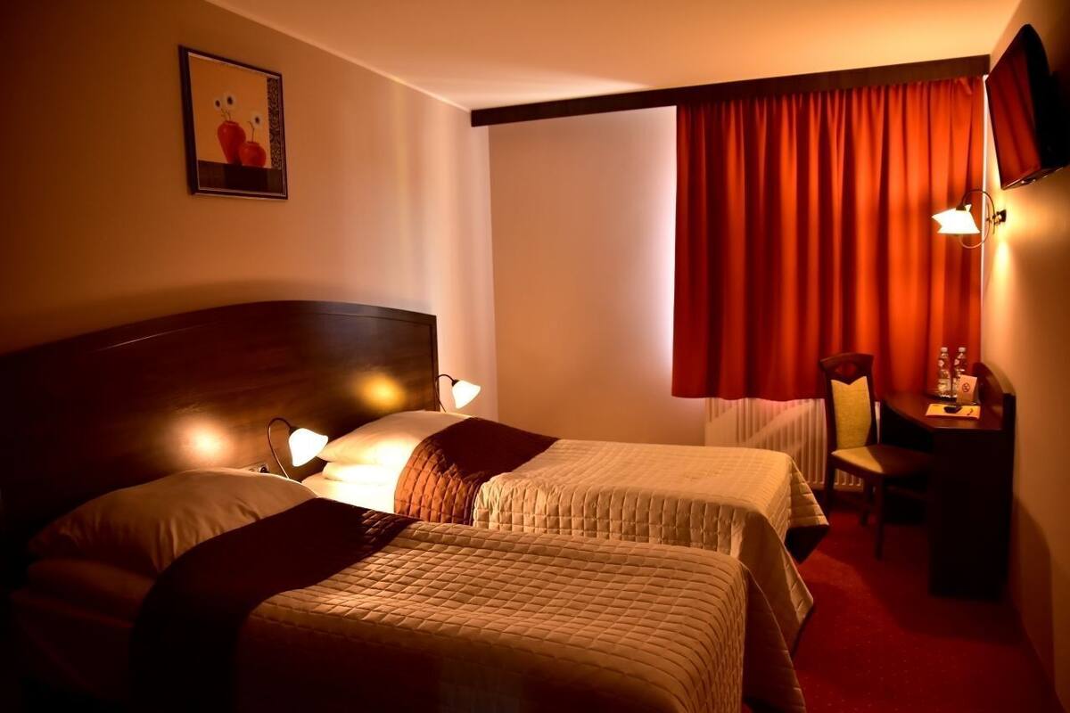 A hotel room with two beds, a desk, a chair, a TV, and a window with red curtains.