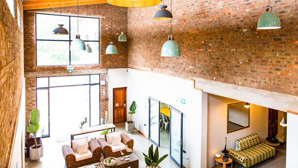 A spacious lobby with a high ceiling, exposed brick walls, and multiple hanging pendant lights. There are seating areas with couches and a coffee table, and large windows providing natural light. - Execustay (Mbombela)