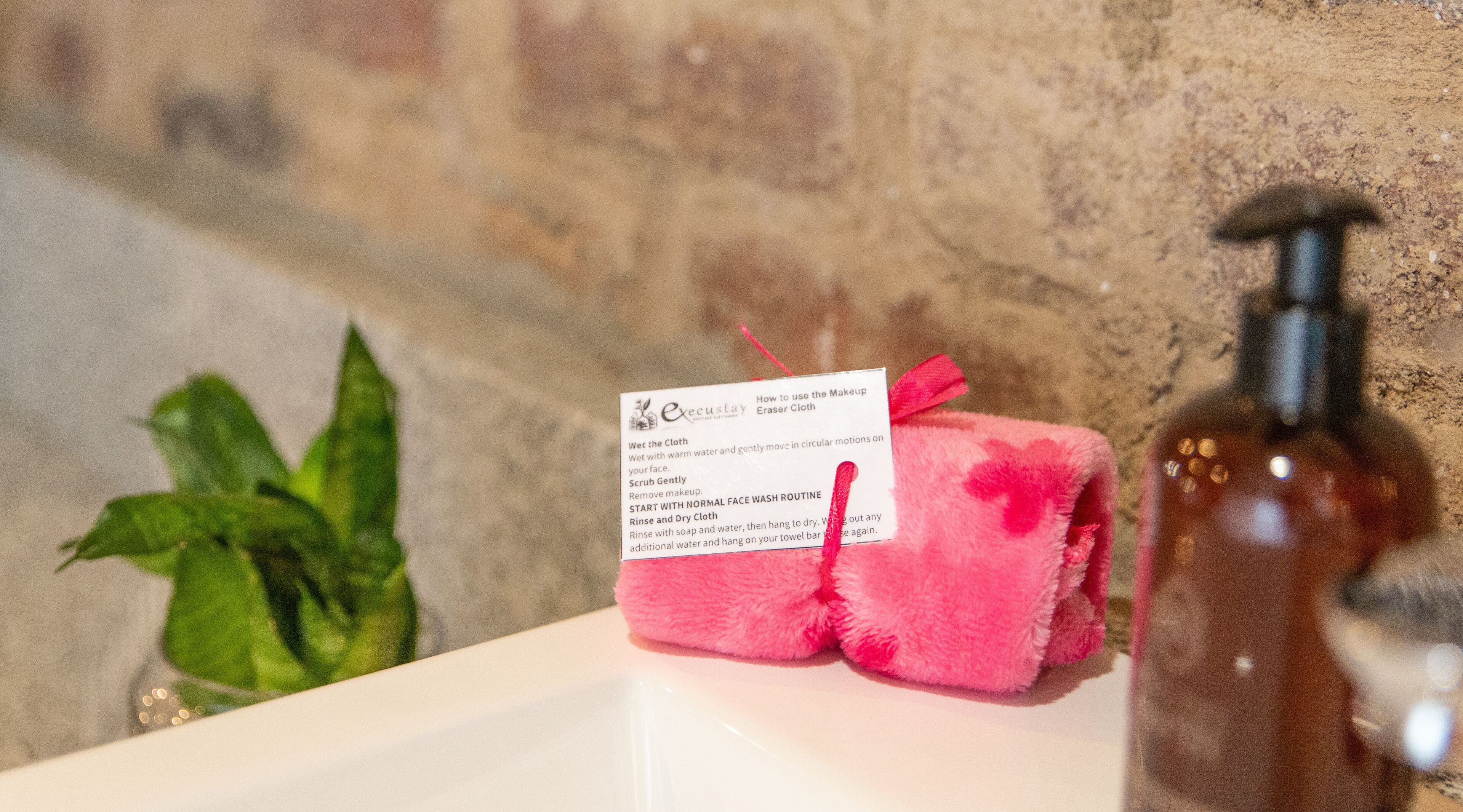 A bathroom with a white sink, a pink towel, a bottle of soap, and a green plant.